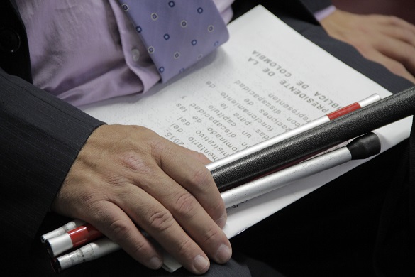 Documento en braile y bastón para personas con discapacidad visual sobre las piernas de un hombre que las sujeta con su mano