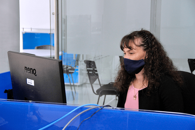 Mujer con tapabocas, sentada y mirando un computador en un puesto de trabajo