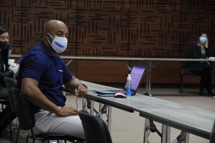 Se ve en primer plano un hombre con tapabocas, sentado en una mesa de conferencias