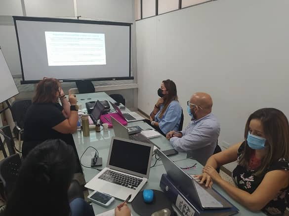 Cuatro mujeres y un hombre, sentados alrededor de una mesa en una sala de juntas, mirando hacia una pantalla en donde se está proyectando algo