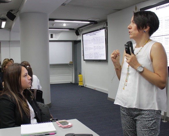 Una mujer de pie exponiendo en un auditorio frente a otra mujer senada