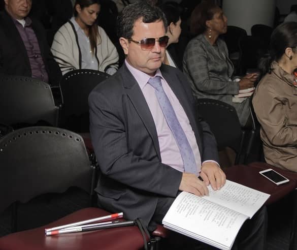 Hombre con discapacidad visual sentado, leyendo un libro en braile que está sobre sus piernas y junto a él, en un silla, hay un bastón