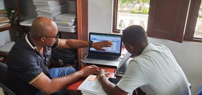 Dos hombres afrodescendientes, sentados uno frente al otro en un escritorio, mirando la pantalla de un computador portátil