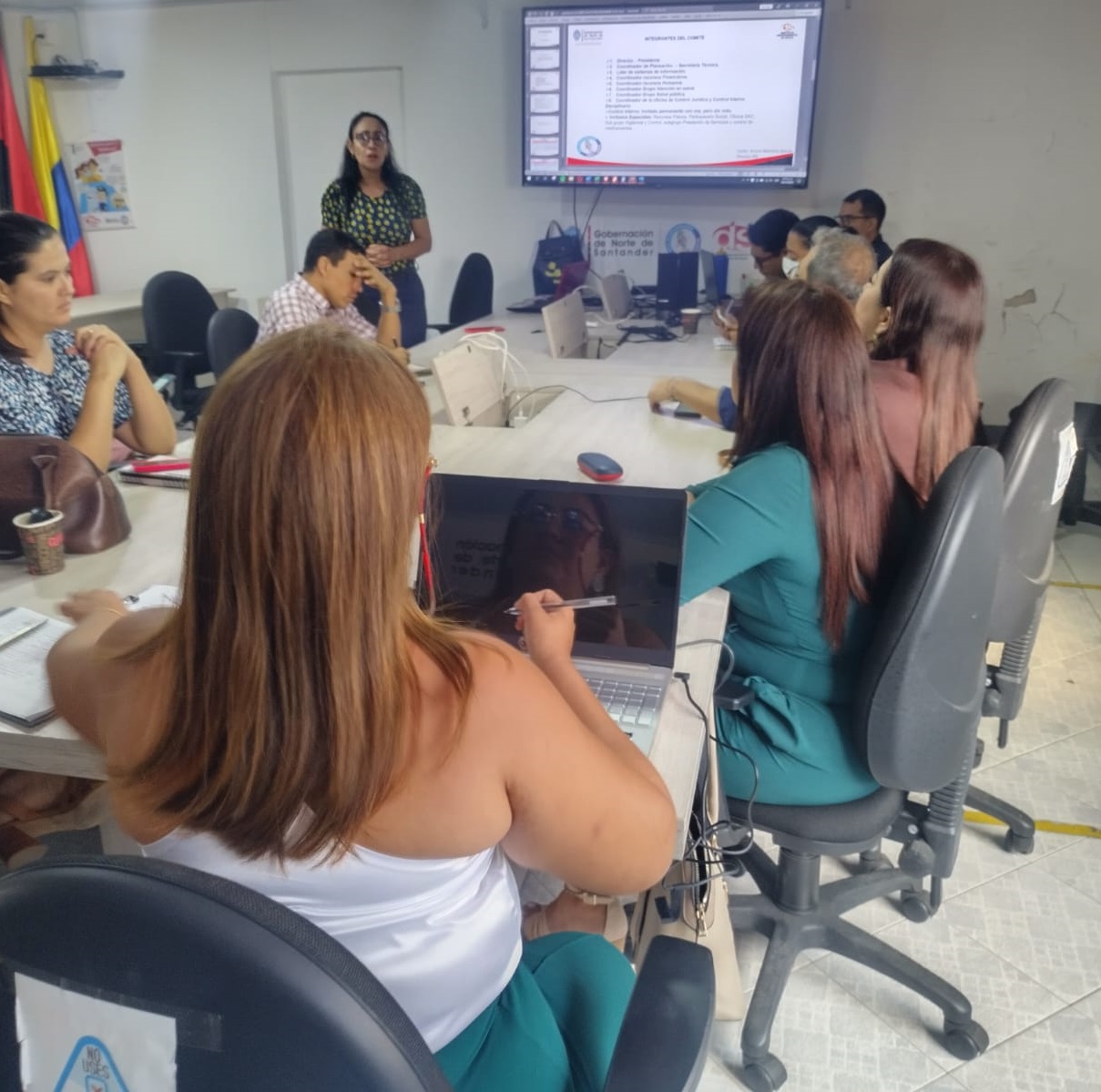 Mujeres y hombres sentados alrededor de una mesa mientras otras de pie expone temas de administración pública 