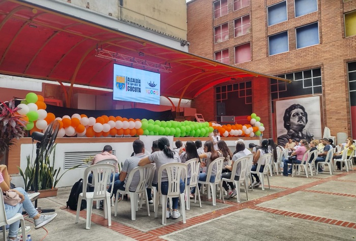 Grupo de personas (hombre y mujeres) sentados frente a una tarima con globos de colores