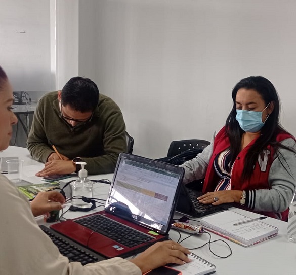 Tres personas sentadas alrededor de una mesa, un hombre escribiendo a mano y dos mujeres, cada una frente a un computador portátil