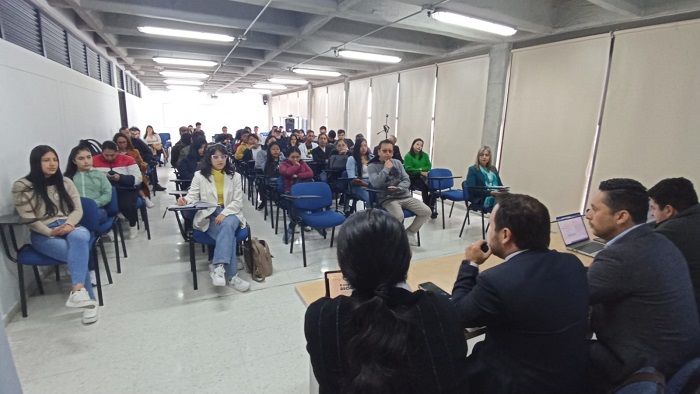 Un aula con un grupo de cuatro personas, tres hombres y una mujer, sentados adelante como expositores en una mesa, y un grupo de personas sentadas en sillas como auditorio