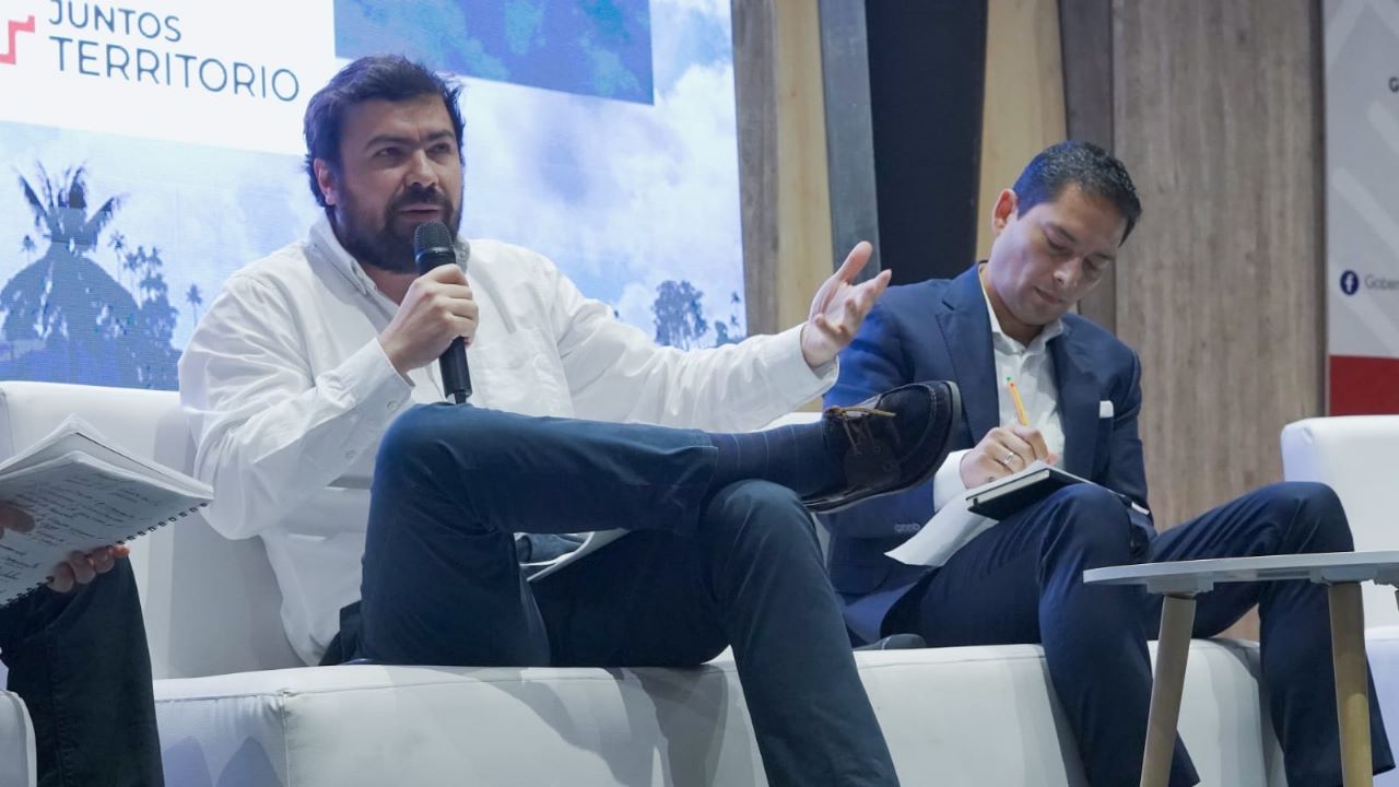 Dos hombres sentados en un sofa, uno hablando por un micrófono y el otro escribiendo sobre un documento que apoya en sus piernas