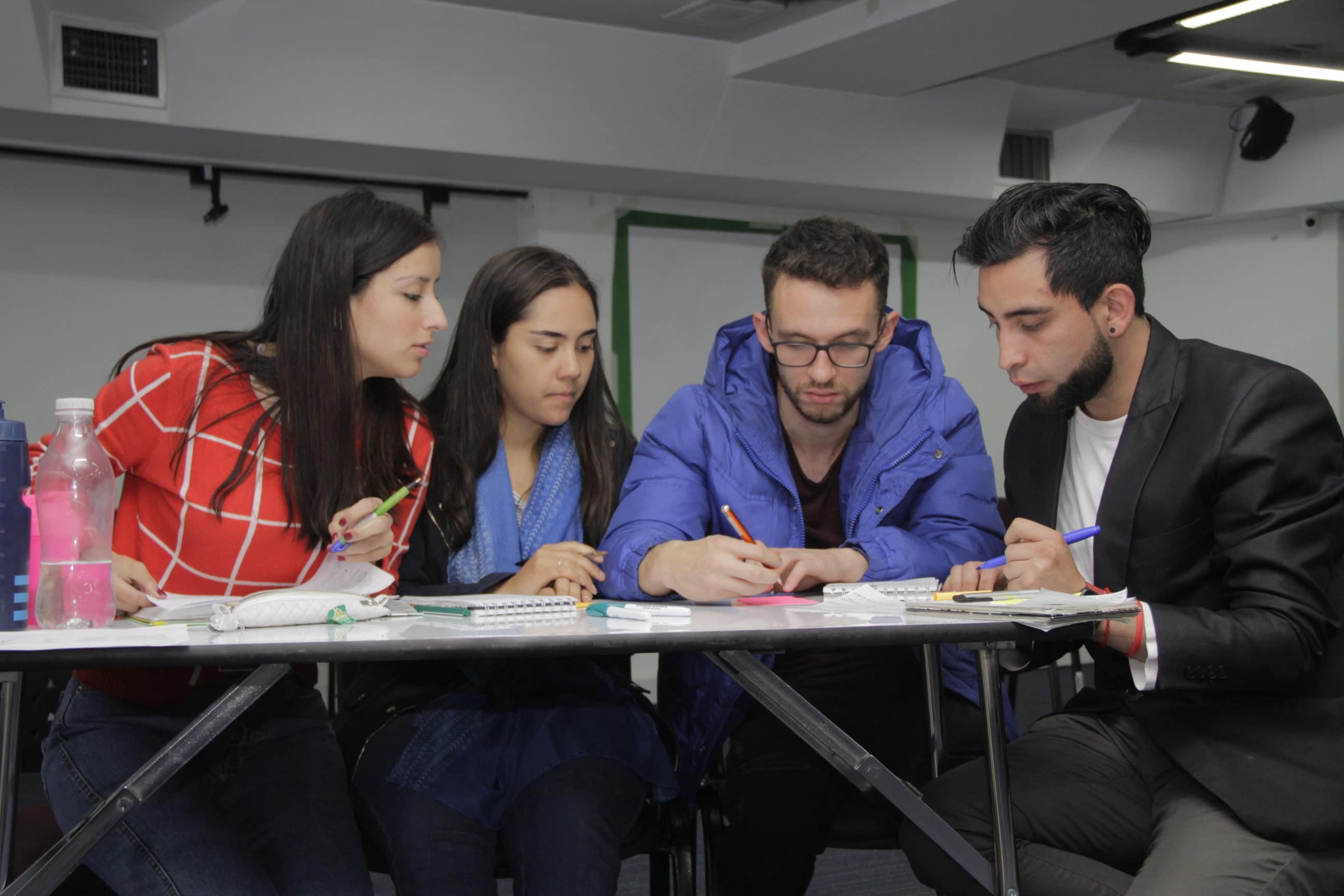 Cuatro jóvenes, dos mujeres y dos hombres, sentados en un escritorio escribiendo en libretas de apuntes. 