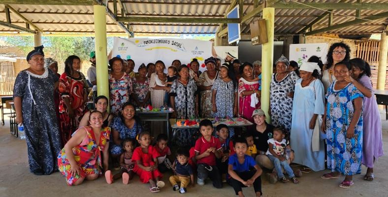 Grupo de mujeres wayúus de pie alrededor de una mesa, la acompañan un grupo de niños