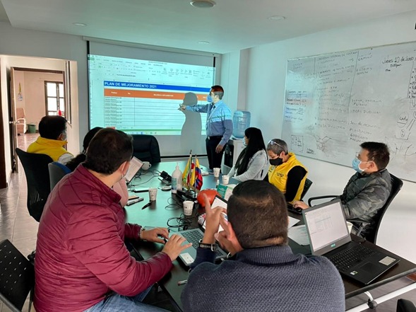 Varias personas en una mesa mirando a un expositor que está frente a una pantalla con estadísticas.