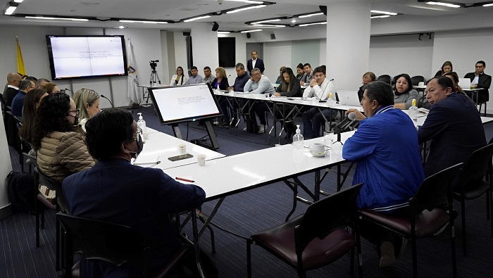 En un auditorio, un grupo de personas reunidas y dentadas al rededor de mesas en forma de mesa redonda