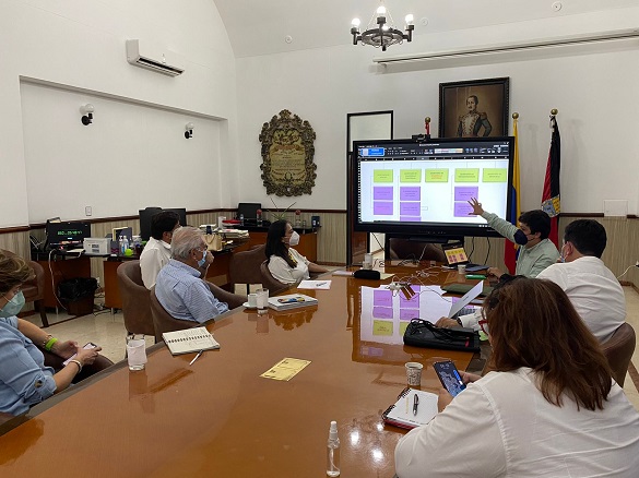 Tres mujeres y cuatro hombres sentados observan una pantalla en una sala de reuniones