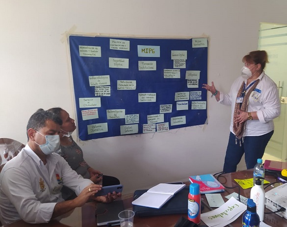 Mujer con tapabocas de pie frente a una cartelera, está explicando o exponiendo ante dos personas, un hombre y una mujer, que se encuentran sentados presando atención