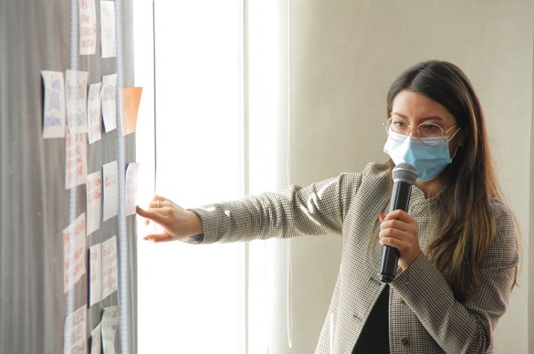 Mujer con tapabocas y micrófono en la mano señalando con su dedo a unos postit en la pared