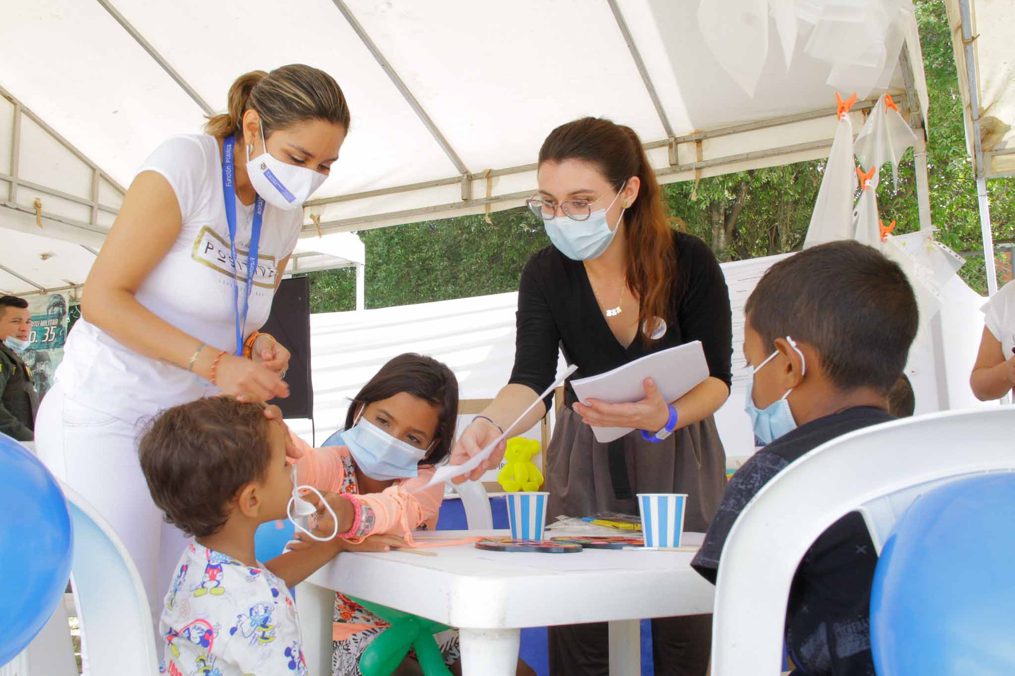 Dos mujeres con tapabocas entregando un documento a tres niños que están sentados alrededor de una mesa